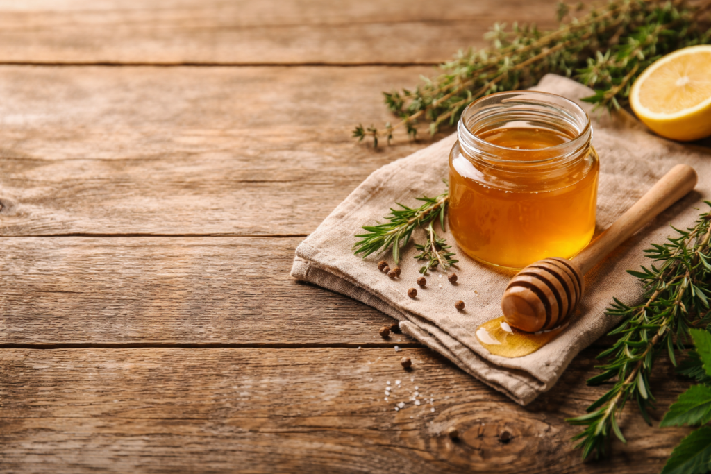 Honigglas und Honiglöffel auf einem Leinentuch. Arrangiert auf einem rustikalem Holztisch dekoriert mit Rosmarin und Thymianzweigen